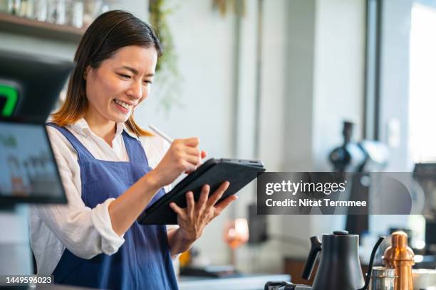 digitization at the point of sale in coffee shop business. female japanese waitress working inside the cashier counter while using a tablet computer for orders received from online customers. - barista coffee restaurant stock pictures, royalty-free photos & images