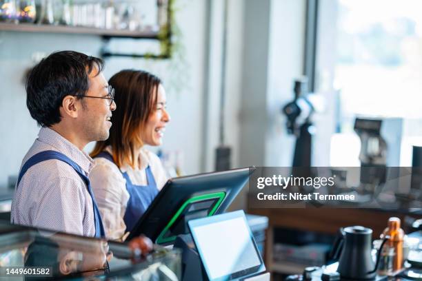 turnkey coffee shop and cafe franchise opportunities. shot of a male japanese coffee shop owner and a female waitress at a cashier counter and greeting customers as they enter the coffee shop. - franchise stock pictures, royalty-free photos & images