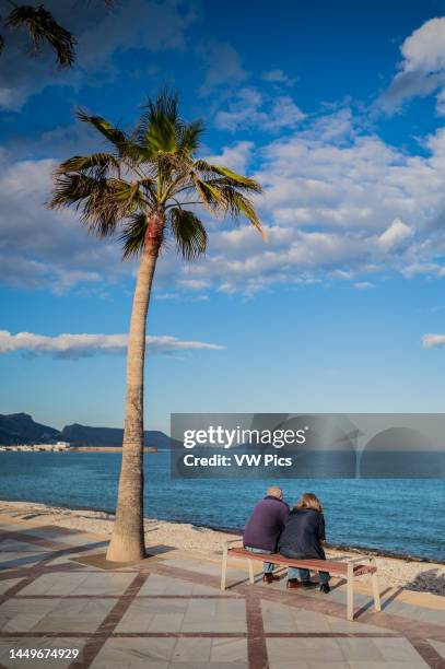 Mature couple sitting in Albir beach promenade at sunset, Alicante, Spain.