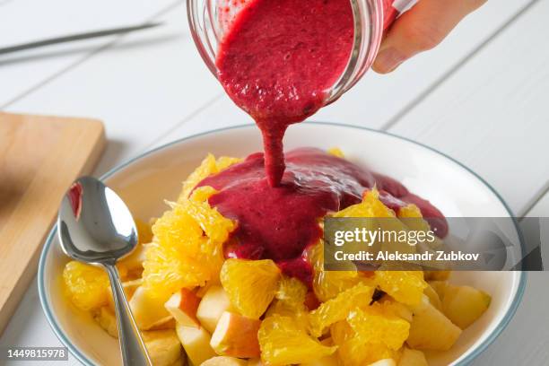 a woman or a girl eats a fruit salad in a ceramic plate, pours a berry blueberry cocktail or yogurt from a glass glass, against the background of a white wooden dining table. healthy food, diet. vegetarian, vegan and raw food. ethical consumption. - pouring softdrink stock-fotos und bilder