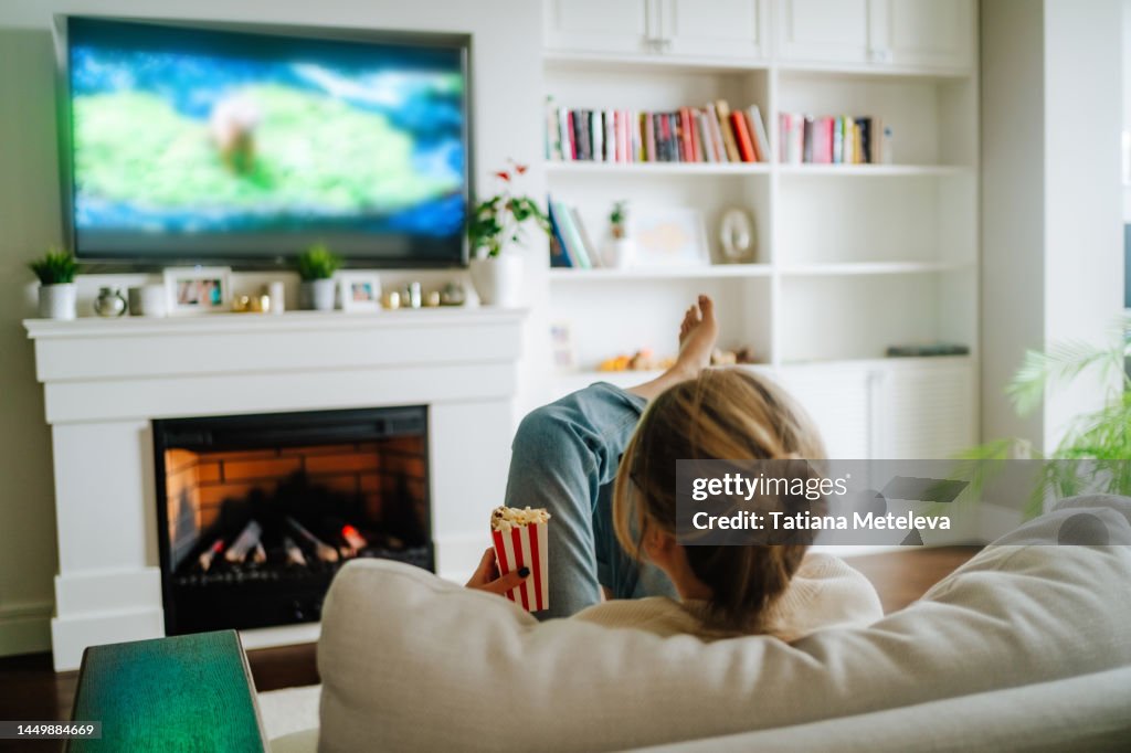 Cozy hugge fireplace and watching TV. Woman eating popcorn and watching tv on a big screen at home
