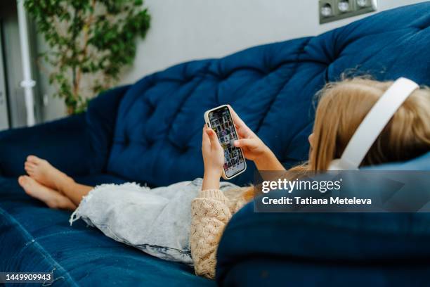 listen to music trends and playing games on a sofa. schoolgirl using smartphone with headphones, lying on a blue sofa in living room - podcast headphones stock pictures, royalty-free photos & images