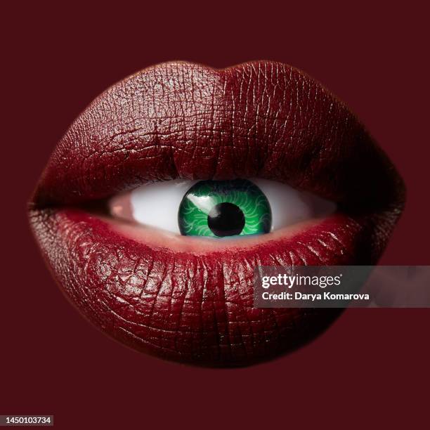 a green eye in the lips on isolated red background, the concept of lip augmentation, too much love for beauty manipulation. an afro american woman’s red lips holds a blue plastic eye. the color of the 2023 year. - women open mouth stock pictures, royalty-free photos & images