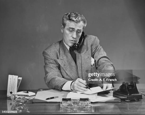 Man wearing a grey suit sitting at a desk, a telephone receiver held in the crook of his neck, as he holds a pen and a document in his hands, United...