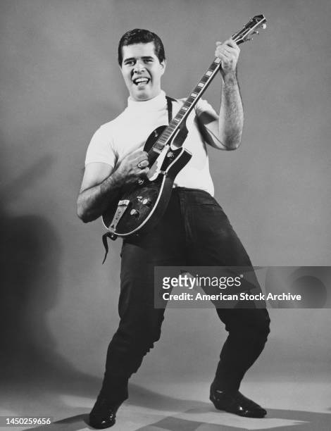 Man wearing a white short-sleeve, turtleneck sweater and black trousers, leaning back as he plays an Gibson electric guitar, United States, circa...