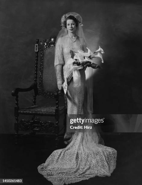 Bride, her halo veil lifted, in a lace wedding dress, the train gathered at her feet, poses with a bouquet of Calla lilies, beside an intricately...