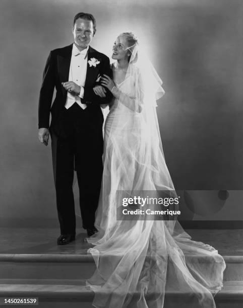 Groom and bride, in a black suit and a wedding dress, the bride's veil lifted, and the train gathered at her feet, as the couple stand smiling...