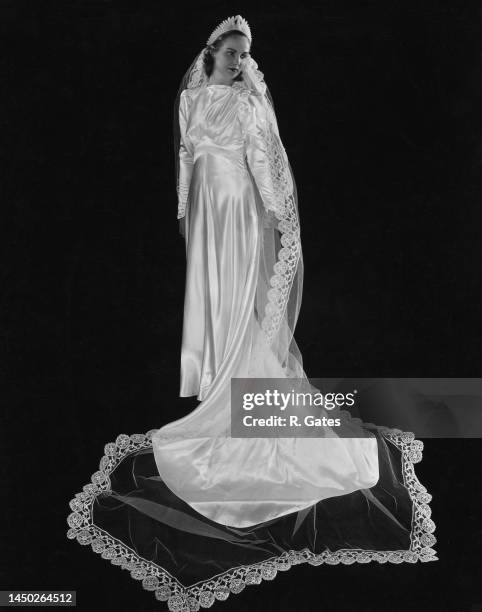Bride wearing a satin wedding gown with a long train gathered at her feet, posing against a black background, United States, circa 1943.