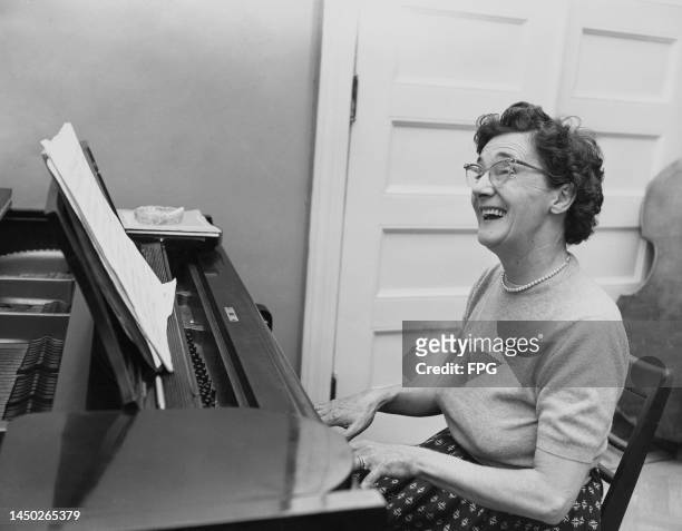 An smiling woman, wearing a short-sleeve crew-neck sweater and a pearl necklace, playing the piano, sheet music propped against a music stand on top...