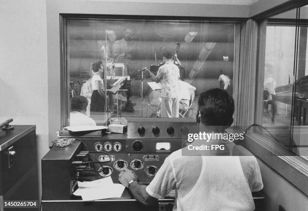 Rear view of an sound engineer sitting in the control booth, looking through a window, with musicians in a recording studio on the other side of the...