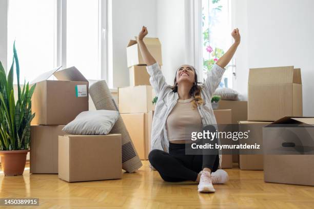 young woman sitting on floor in new apartment with boxes and raising arms in joy - student loan stockfoto's en -beelden