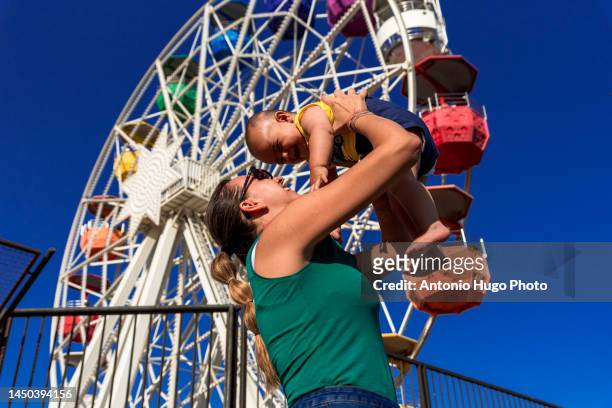 mother lifting her baby in her arms. colorful ferris wheel in the background at an amusement park. - famous family funfair stockfoto's en -beelden