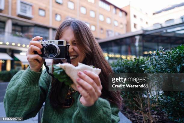 happy woman holding sandwich photographing through camera in front of building - in front of camera stock pictures, royalty-free photos & images