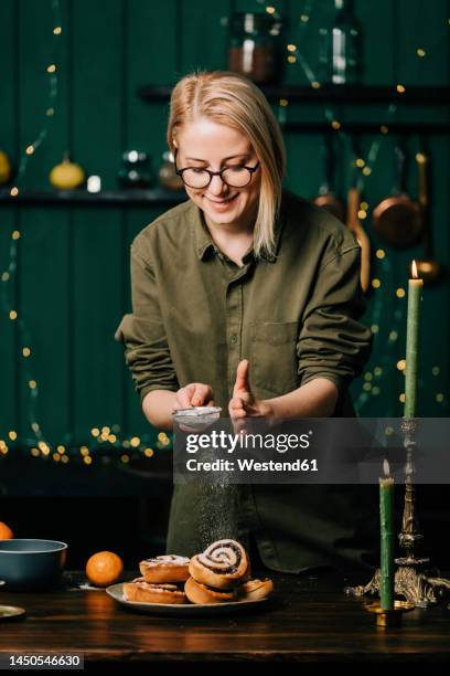 smiling woman sprinkling powdered sugar on cinnamon buns in kitchen - powdered sugar stock-fotos und bilder