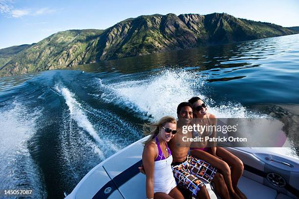 three young adults laugh and smile while sitting on the back of a wakeboard boat on a lake in idaho. - pend orielle lake stock pictures, royalty-free photos & images