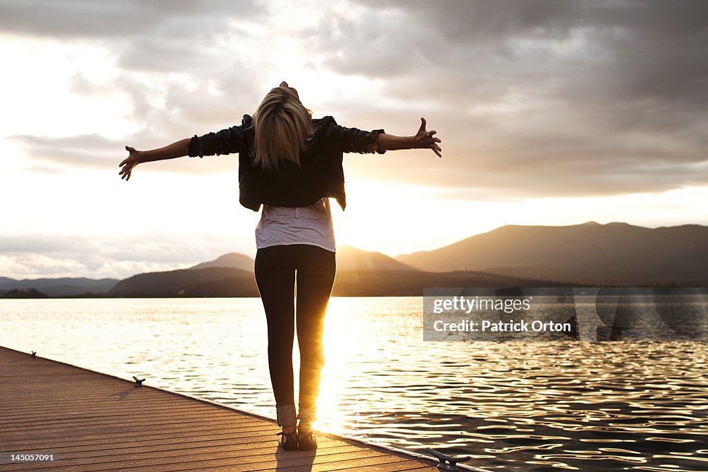 A beautiful young woman looking into the distance holds her arms out embracing the sunset over a lake in Idaho.