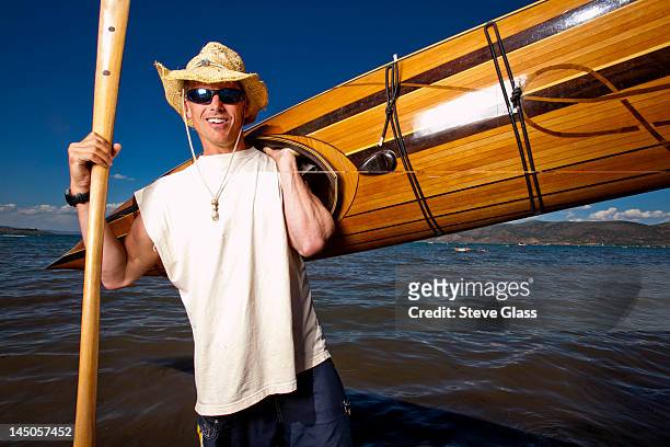a man wears a straw hat as he poses for a portrait with a wooden kayak and paddle that he made while standing on the shore of bear lake, utah. - carrying canoe stock pictures, royalty-free photos & images