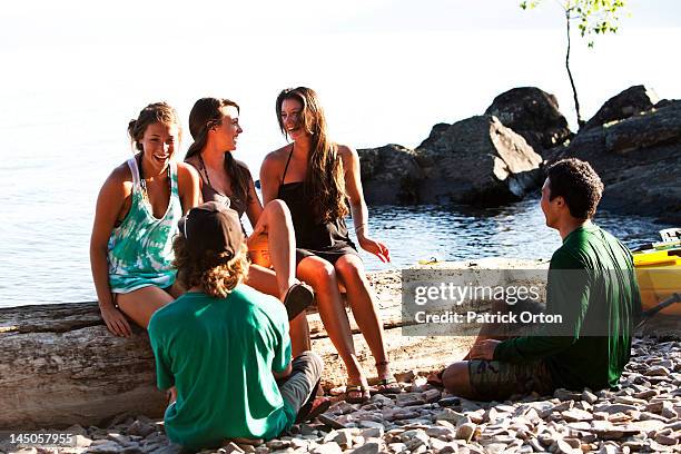 a group of friends laugh and smile sitting on a log next to a lake on a camping trip in idaho. - pend orielle lake stock pictures, royalty-free photos & images