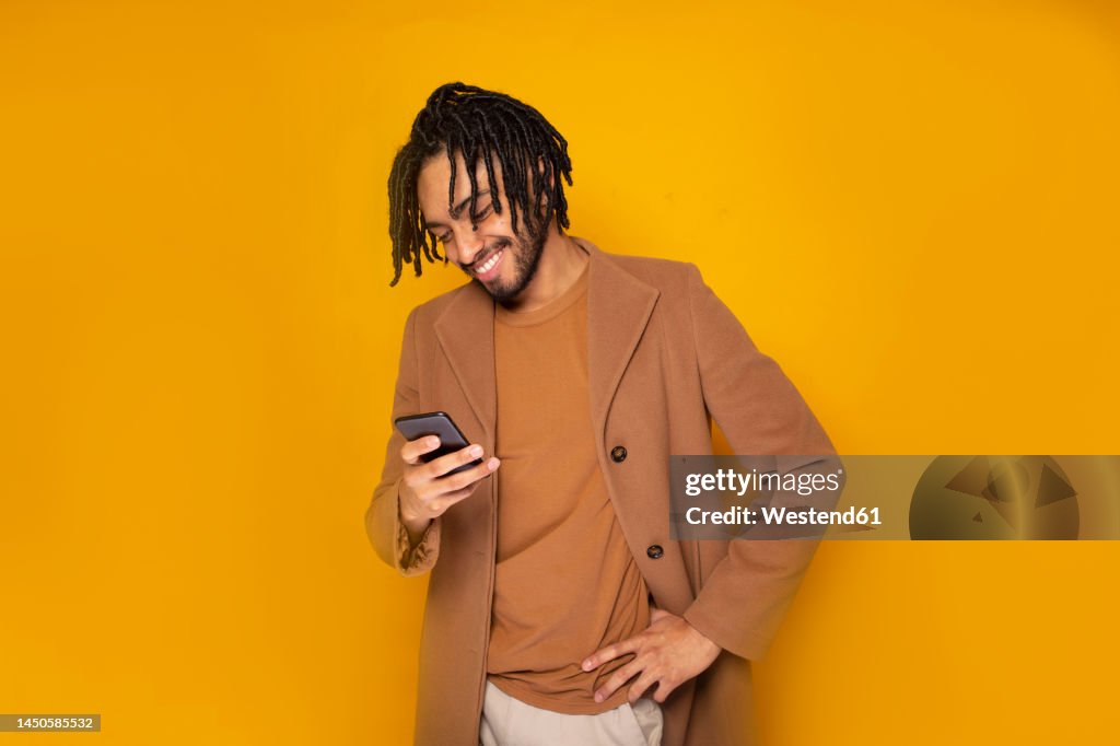 Smiling man with hand on hip using smart phone against yellow background