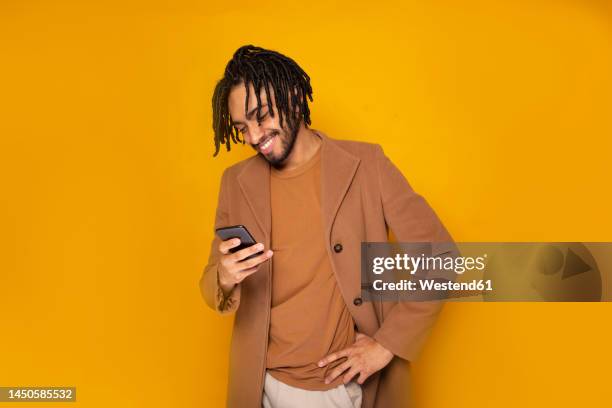 smiling man with hand on hip using smart phone against yellow background - phone coloured background stock pictures, royalty-free photos & images