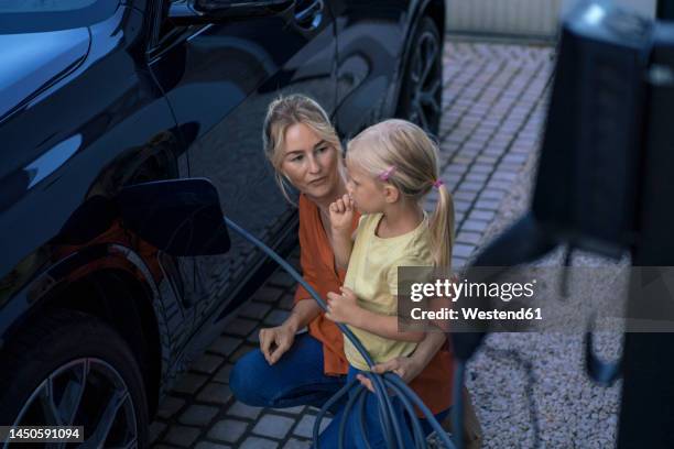 girl with mother charging electric vehicle in front yard - car yard stock-fotos und bilder