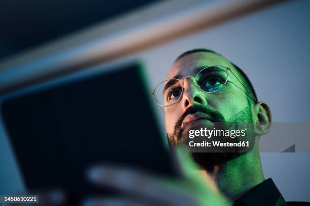 green light falling on thoughtful businessman holding tablet pc - mann mitte 30 stock-fotos und bilder