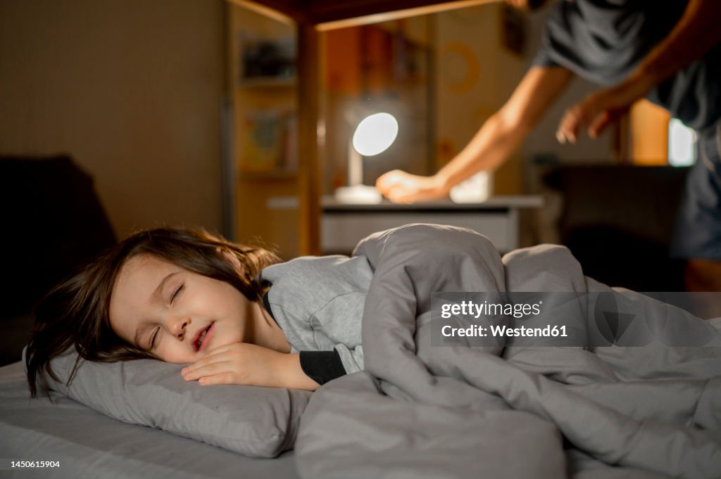 Son sleeping on bed with father putting lights off in background