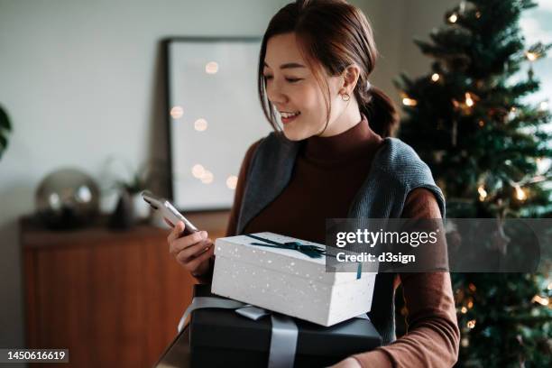 beautiful smiling young asian woman using smartphone while holding wrapped christmas presents, standing against decorated christmas tree. christmas lifestyle. christmas shopping. receiving christmas gifts from friends and family. holiday and festive vibes - christmas shopping stock pictures, royalty-free photos & images