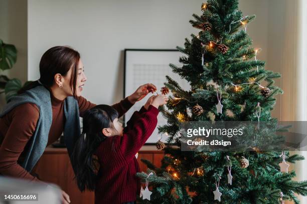 young asian mother and lovely little daughter decorating christmas tree at home together. christmas lifestyle theme. celebrating christmas. holiday and festive vibes. christmas is here - xmas together stock-fotos und bilder