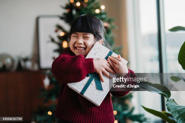 happy little asian girl hugging a box of christmas present on christmas morning, showing her excitement. standing in front of christmas tree in the living room at home. christmas lifestyle theme. celebrating christmas. holiday and festive vibes - kerst cadeau meisje stockfoto's en -beelden