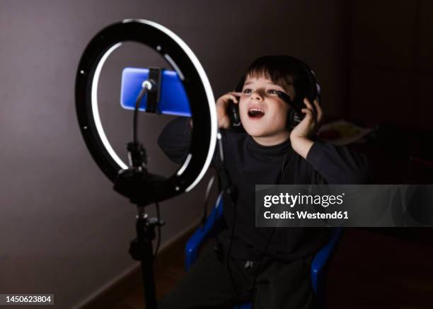 boy wearing headphones recording video through mobile phone in illuminated ring light - live broadcast stock-fotos und bilder