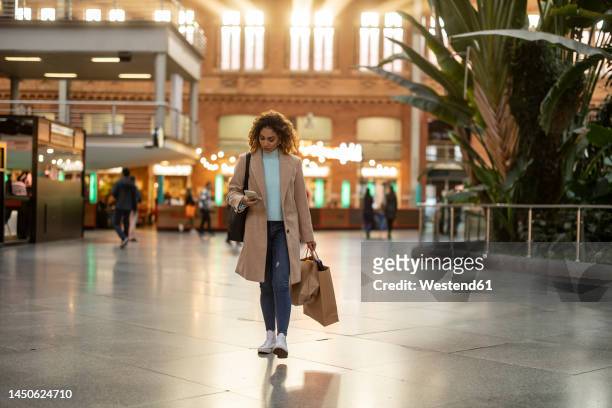 young woman holding shopping bags using mobile phone walking at station - überzieher stock-fotos und bilder
