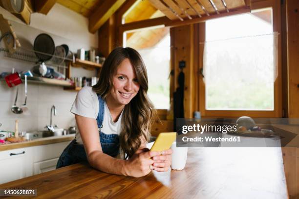 smiling young woman leaning with smart phone on kitchen island - baita interno foto e immagini stock