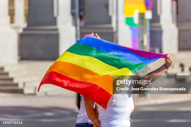 rear view of proud couple of young women attending the gay pride parade in madrid showing rainbow flag through the streets of madrid - homophobia stock pictures, royalty-free photos & images