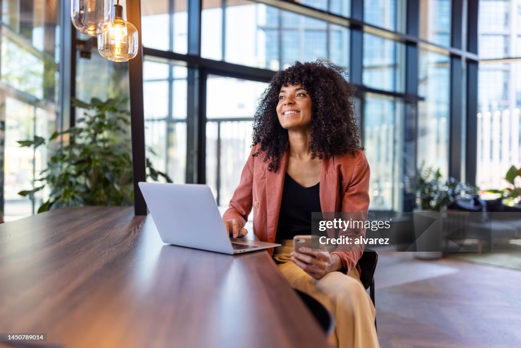 Happy african woman with laptop sitting at table in office lobby