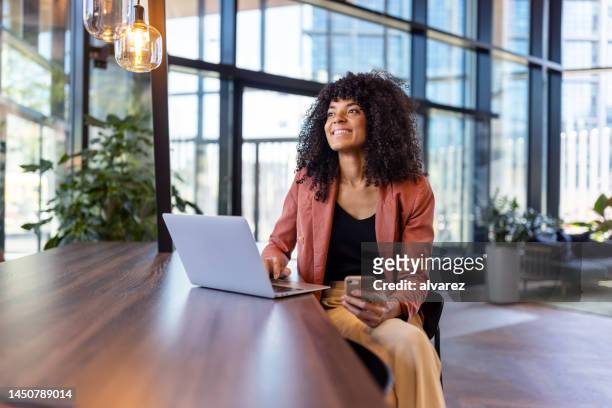 happy african woman with laptop sitting at table in office lobby - facebook founder imagens e fotografias de stock