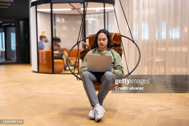 junger mann am laptop sitzt auf einem schaukelstuhl im coworking space - coworking office stock-fotos und bilder