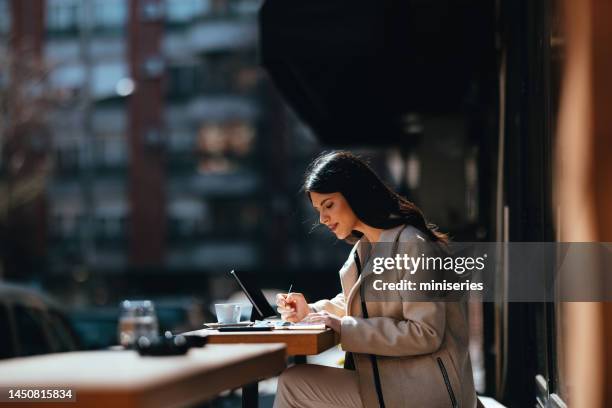 Beautiful Young Businesswoman Sitting at the Coffee Shop Table on the Street and Writing in Her Notebook