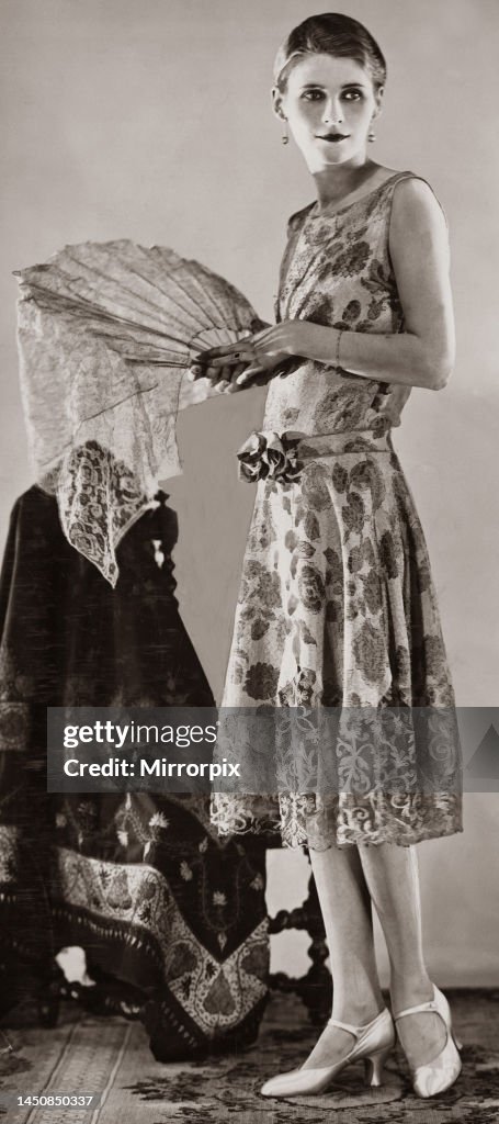 A woman wearing white and gold brocade evening gown which has a tight bodice and narrow belt and bow in front 1926
