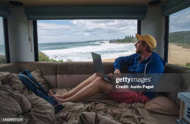 mature man catching up on a bit of work from the back of his motorhome. the view out of the window is of waves rolling into a beach - teleworking stock pictures, royalty-free photos & images