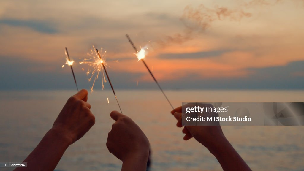 Close up hand of young Asian women celebrating holding sparklers on tropical beach at sunset.