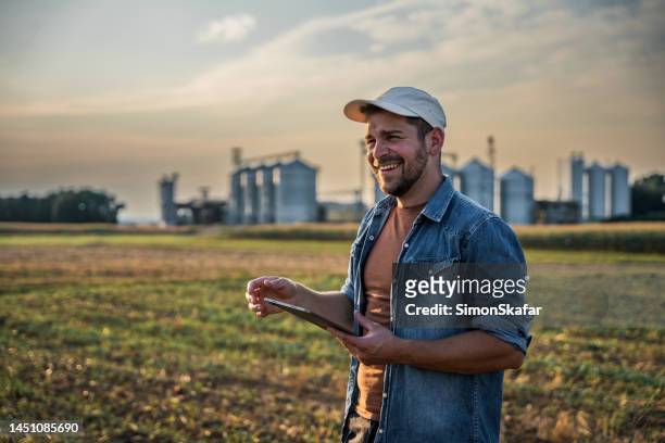 happy male farmer using digital tablet in field against sky - farmer stockfoto's en -beelden