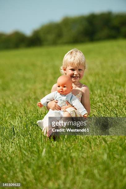 germany, bavaria, girl with baby doll in grass meadow, smiling, portrait - bavaria girl stockfoto's en -beelden