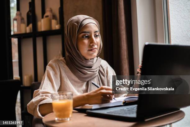 portrait of cheerful muslim woman working online on laptop and communicating by smart phone. - e learning stock pictures, royalty-free photos & images