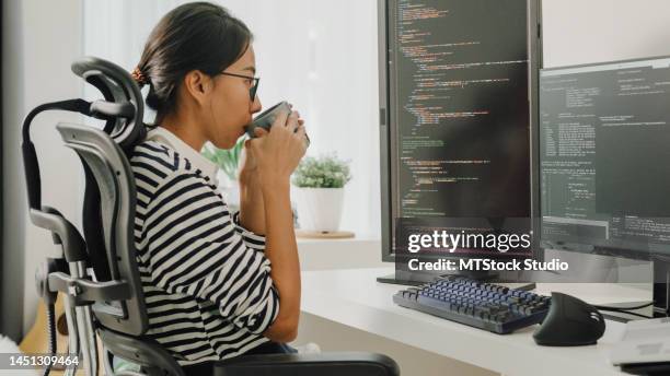 young asian woman software developers drinking coffee and using computer to write code sitting at desk with multiple screens work remotely at home. - java code stock pictures, royalty-free photos & images