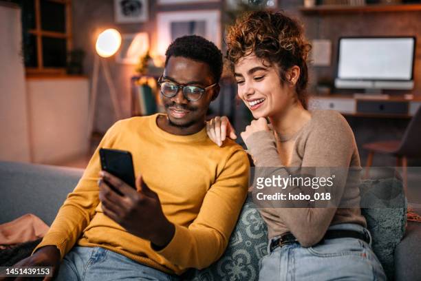casal jovem sentado no sofá, navegando na rede usando o telefone inteligente - smart casual - fotografias e filmes do acervo