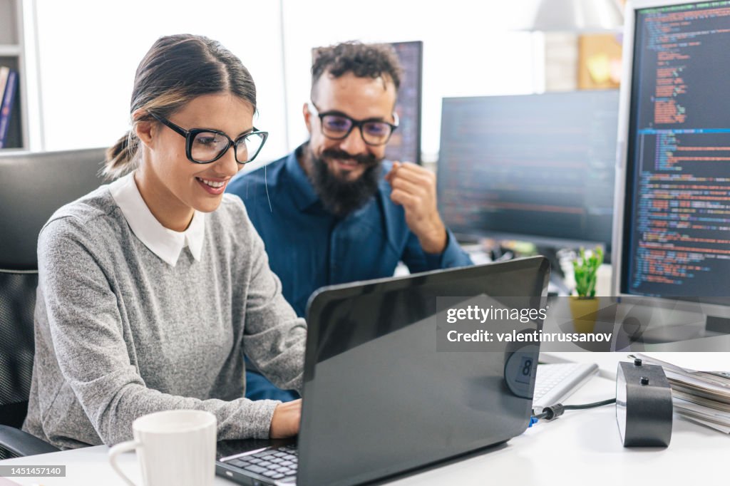 Junge Programmiererin steht auf ihrem Schreibtisch, programmiert auf einem Laptop, diskutiert mit einem männlichen Kollegen, gibt ihr Ratschläge