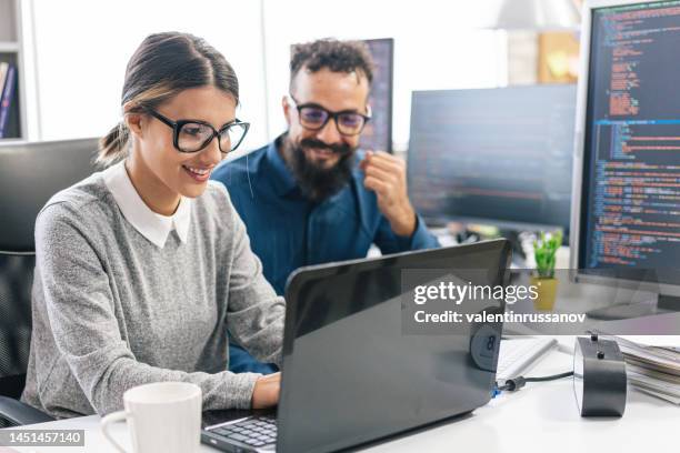 young female programmer standing on her desk, coding on a laptop, having discussion with a male colleague, giving her advice - information security imagens e fotografias de stock