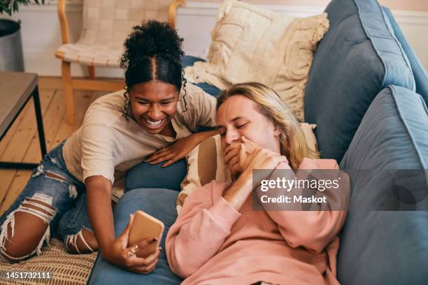 happy teenage girl sharing smart phone with transgender person lying on sofa at home - tears happiness stock pictures, royalty-free photos & images