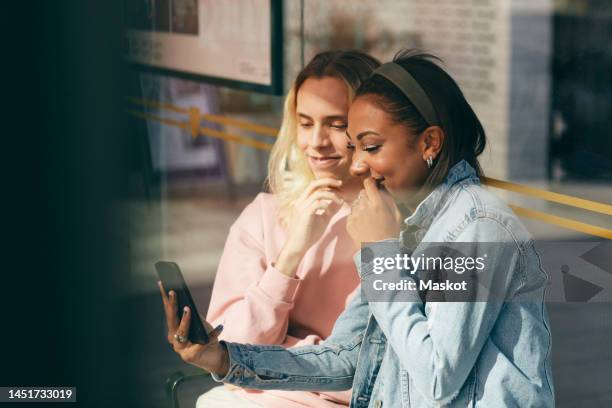 smiling teenage girl sharing smart phone with transgender person sitting near glass wall - girl looking through window stock pictures, royalty-free photos & images